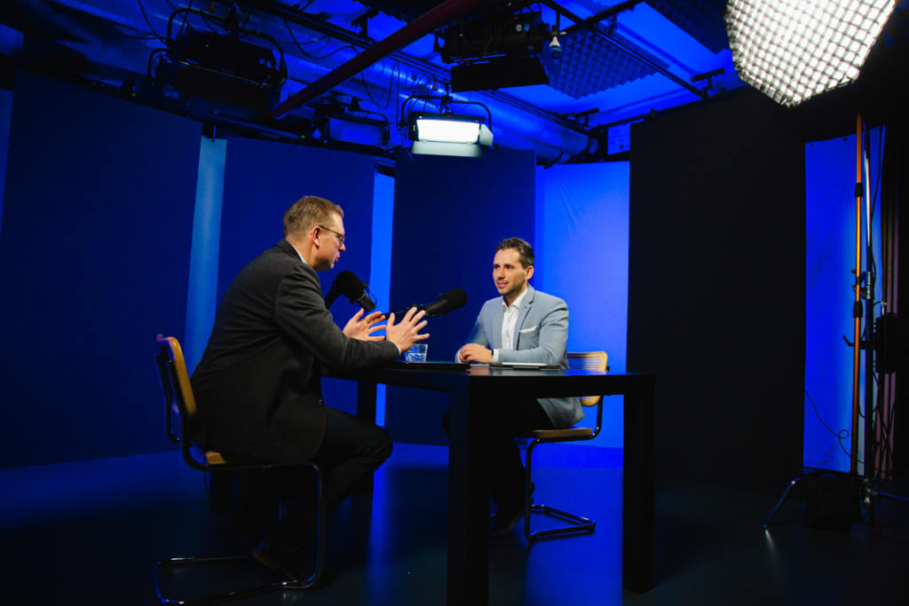 Das Bild zeigt Daniel Arndt (Abteilungsleiter Investment) und Maximilian Kreitlmeier (Teamleiter Portfoliomanagement und Analyse) in einem Video-Podcast Setting. Beide sitzen an einem Tisch mit Mikrofonen und diskutieren miteinander.