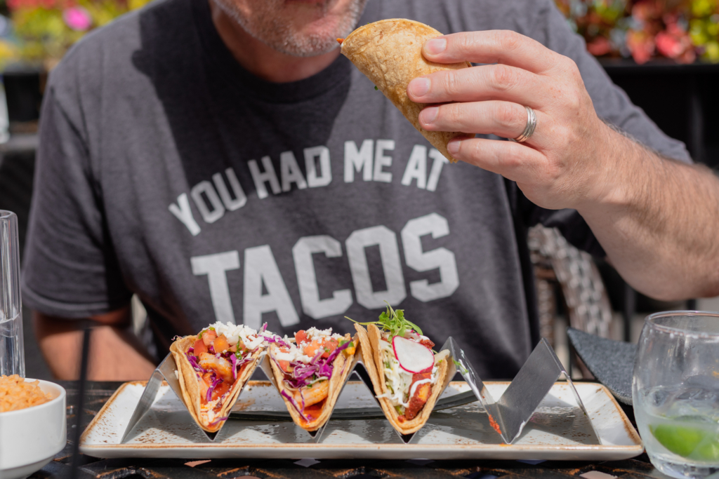 Mann der ein Taco isst. Vor ihm auf dem Tisch steht ein Teller mit drei weiteren Tacos. Auf seinem T-Shirt kann man einen Spruch lesen: You had me at Tacos.